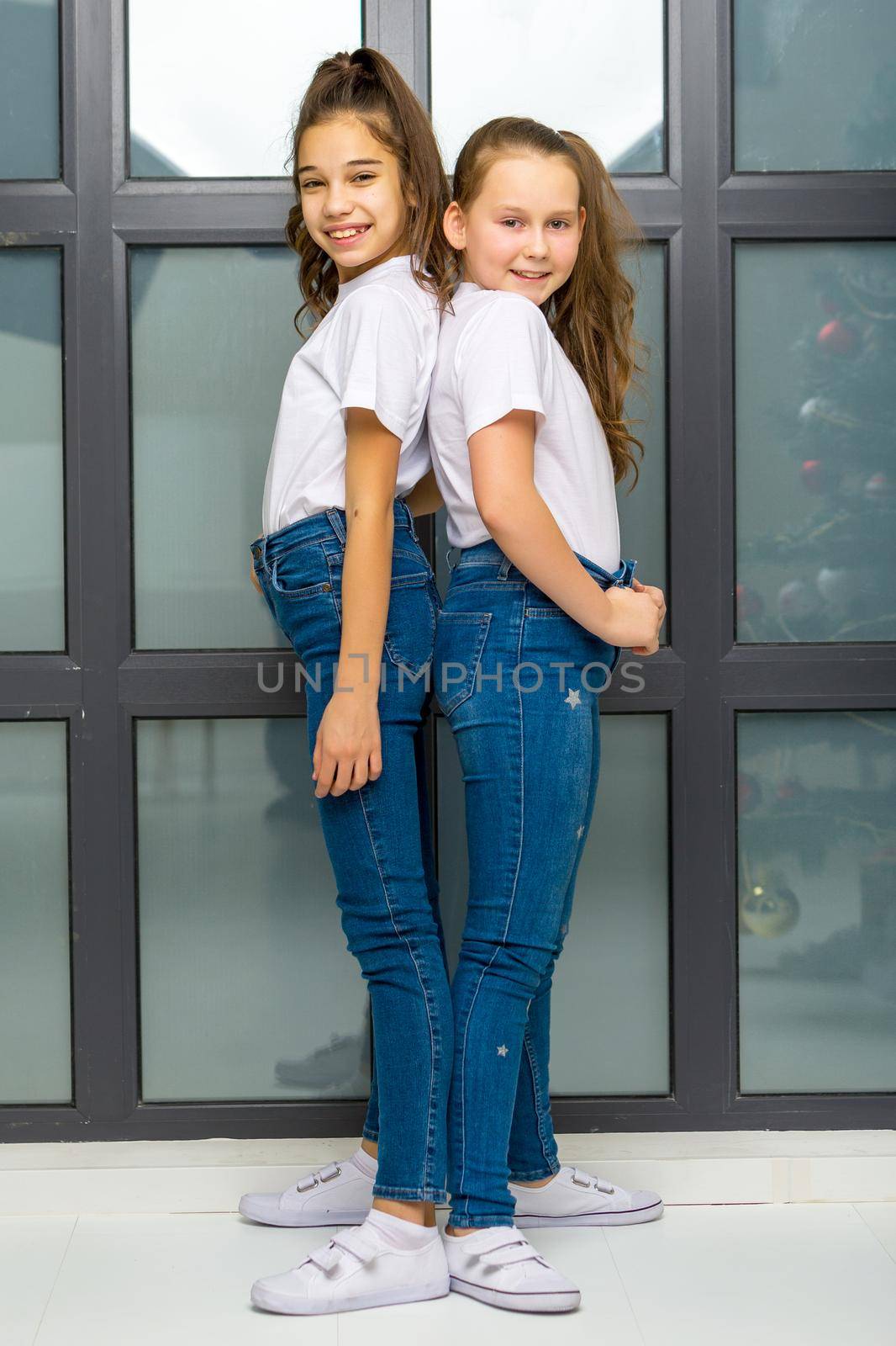 Two beautiful little girls of school age posing in the studio near a large panoramic window. Outside the window it is already evening, dark, the concept of tenderness and beauty.