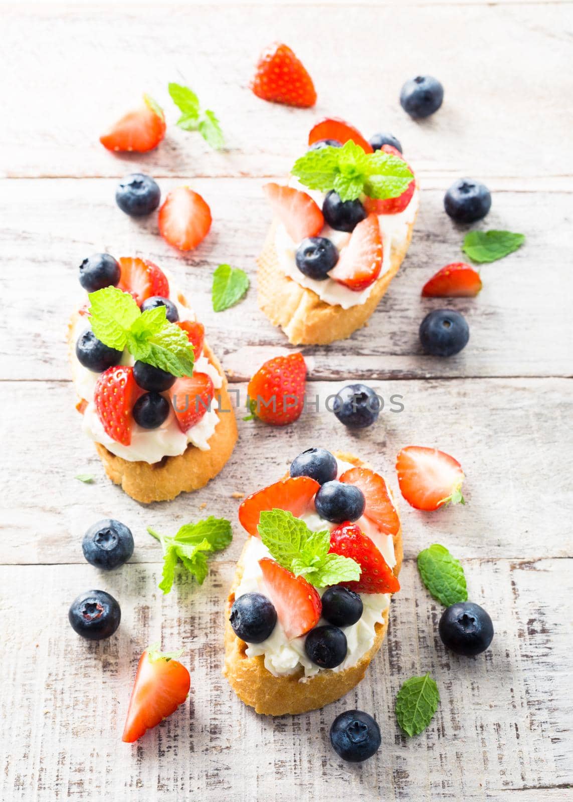 Small canape, crostini with grilled baguette with cream cheese, blueberry, strawberry and mint on old white wooden background. Delicious appetizer or dessert.