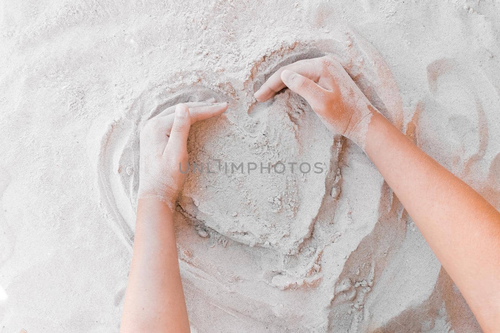 The hand of a young girl draws a heart on the white beach sand close-up. Symbol or sign of love concept.