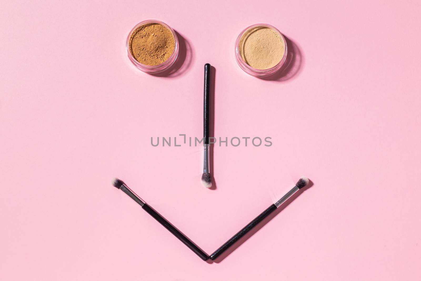 Funny face with make-up brushes and mineral powder on pink background with copy space, top view. Cosmetics humor and beauty concept. by Satura86