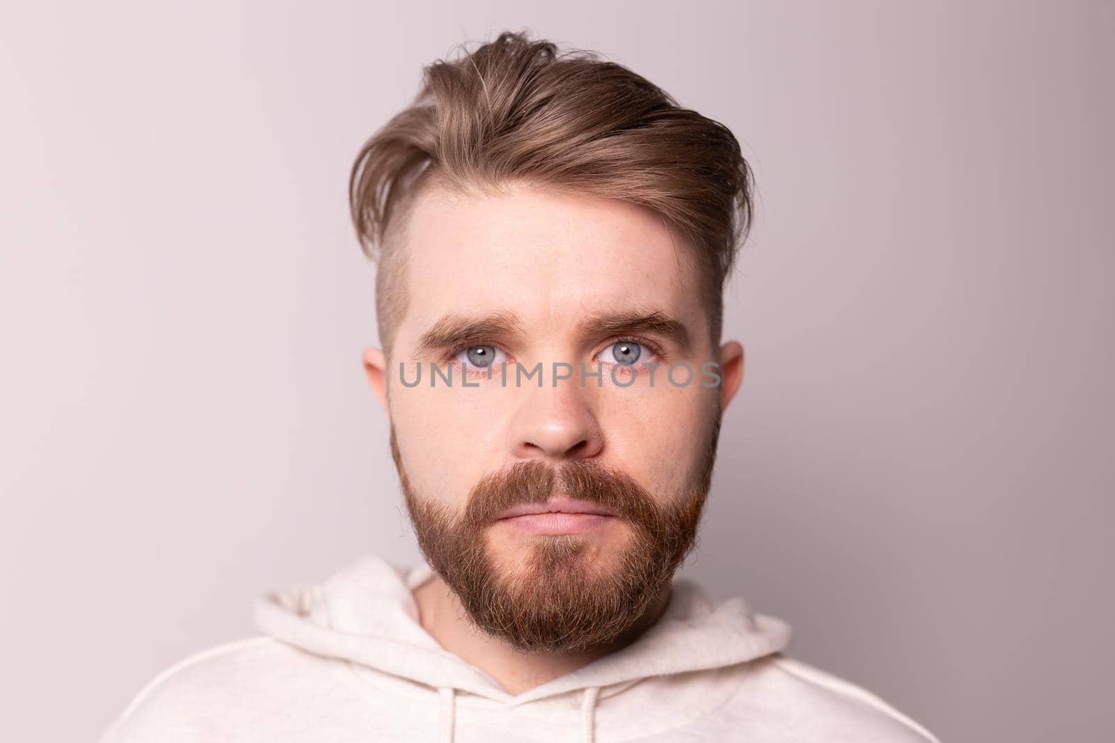 Portrait of young male with beard and mustache and trendy hairdo. Wears casual hoodie, has serious expression in studio against white