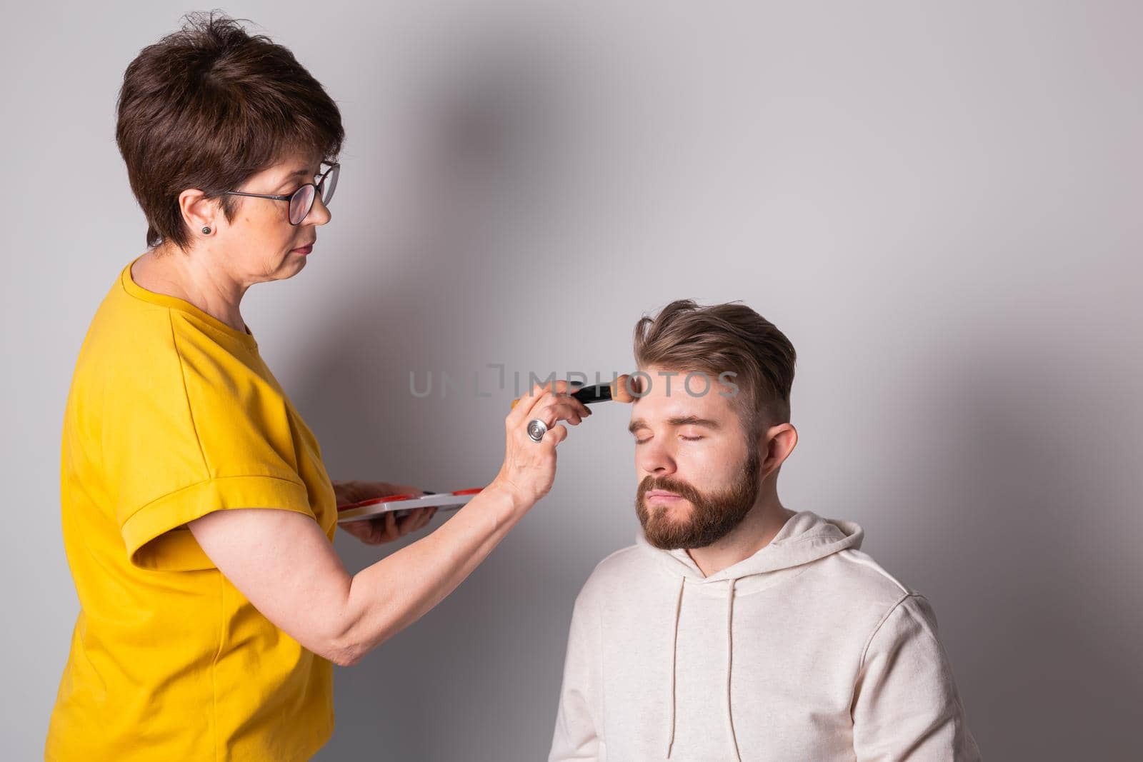 Bearded man getting makeup. Woman visagist works with brush