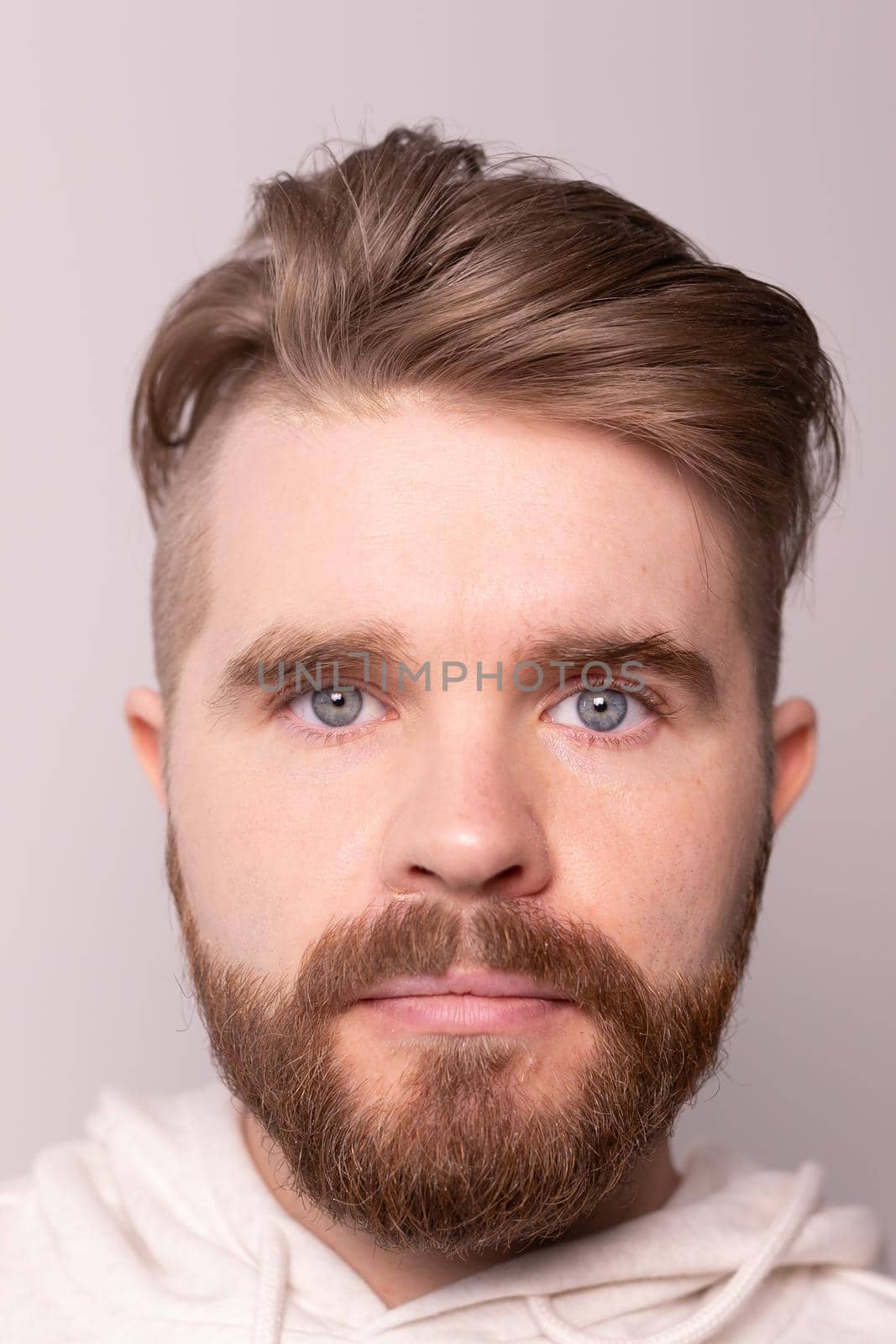 Portrait of young male with beard and mustache and trendy hairdo. Wears casual hoodie, has serious expression in studio against white