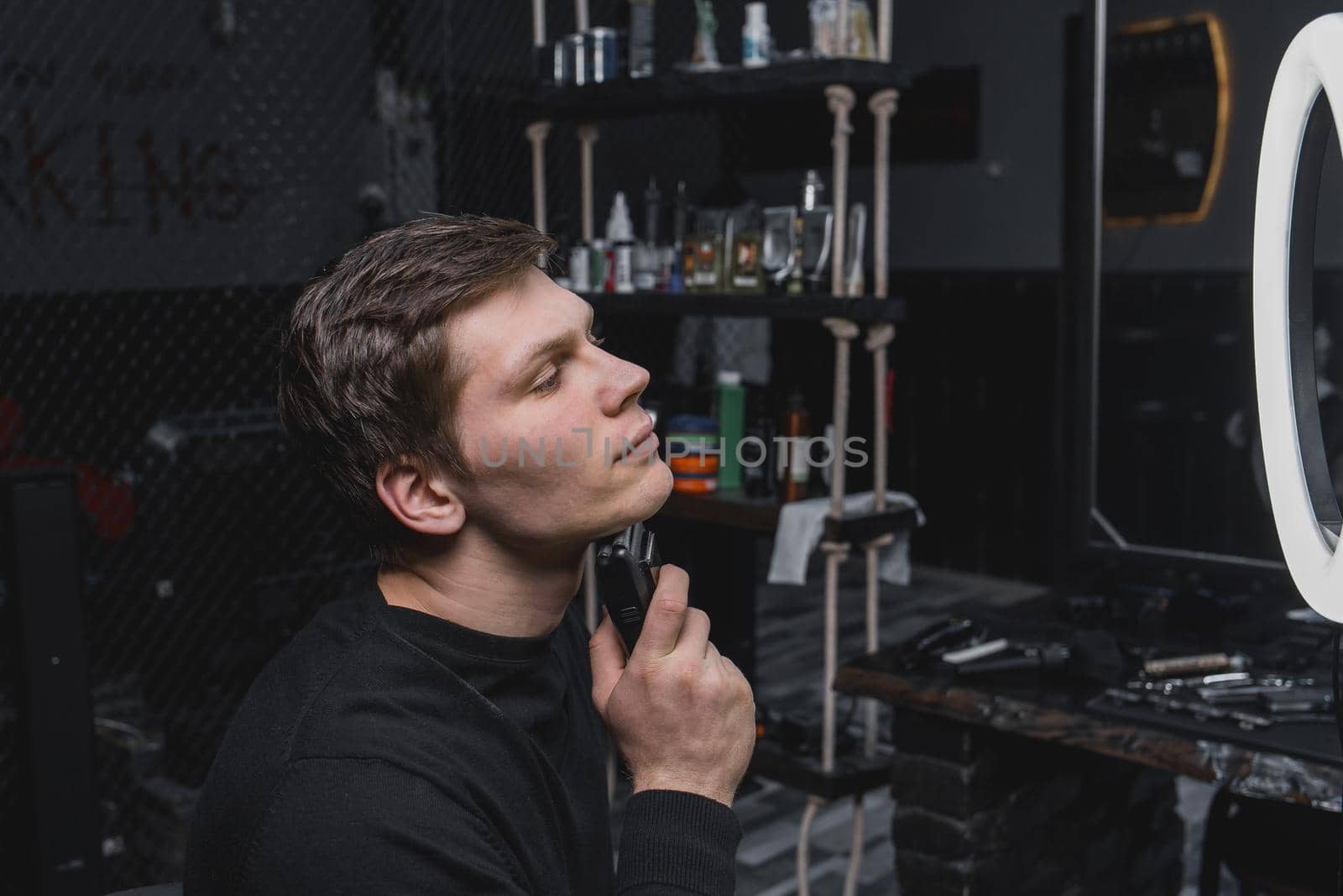 A serious young guy of Caucasian appearance with dark hair shaves with an automatic razor next to the ring lighting. Care for your appearance.