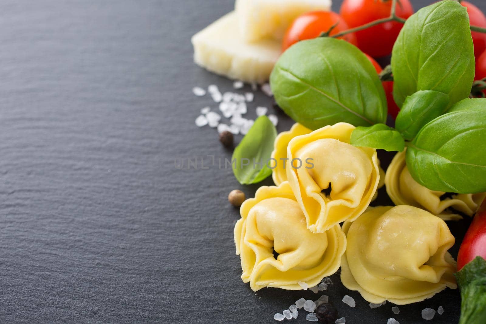 Homemade raw Italian tortellini filled with ham and cheese with raw tomatoes, basil leaves and cheese on black background. Healthy food concept with copy space concept. Selective focus.