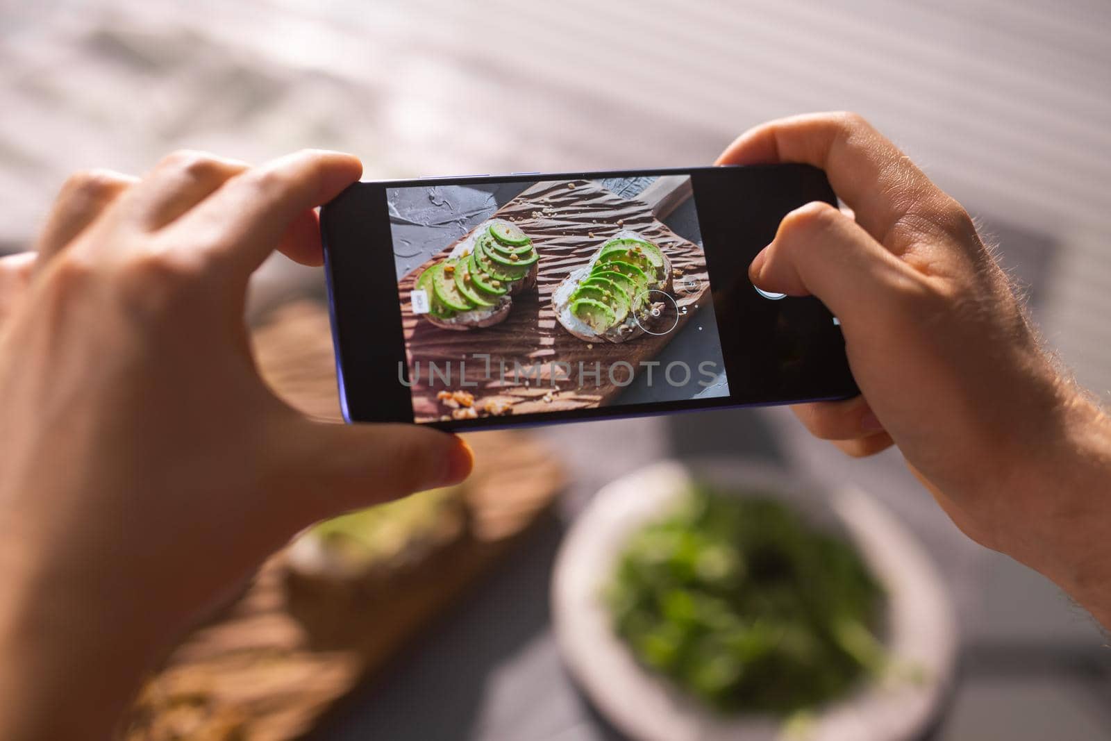 Hands take pictures on smartphone of two beautiful healthy sour cream and avocado sandwiches lying on board on the table. Social media and food