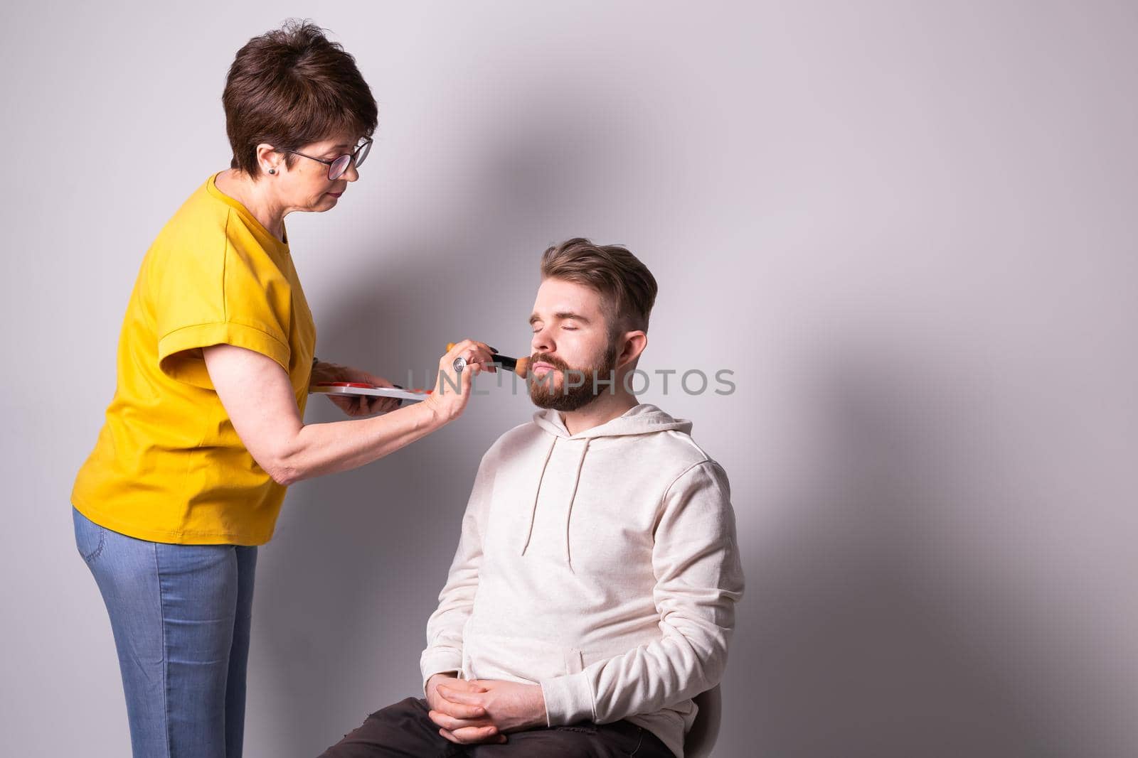 Bearded man getting makeup. Woman visagist works with brush