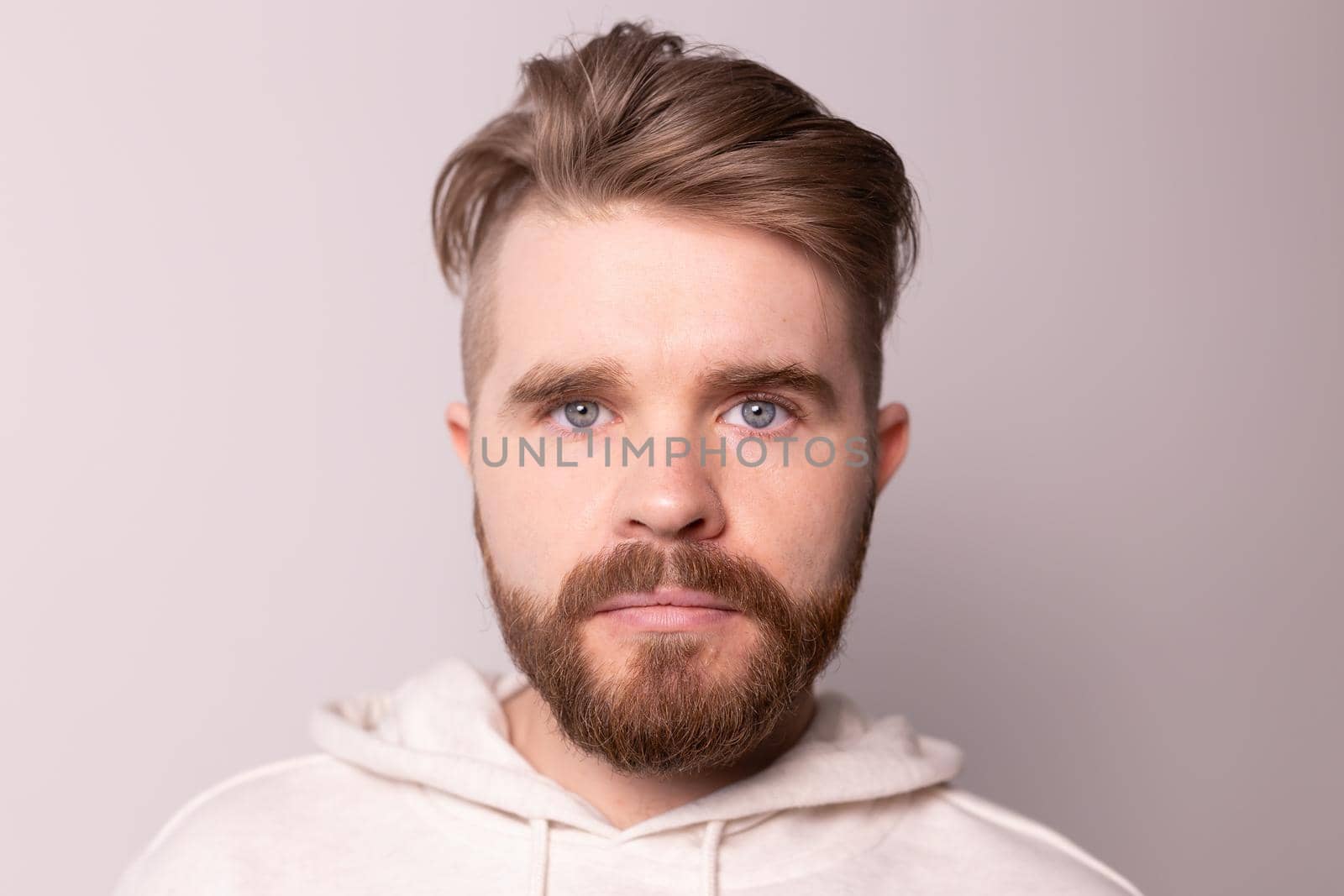 Portrait of young male with beard and mustache and trendy hairdo. Wears casual hoodie, has serious expression in studio against white