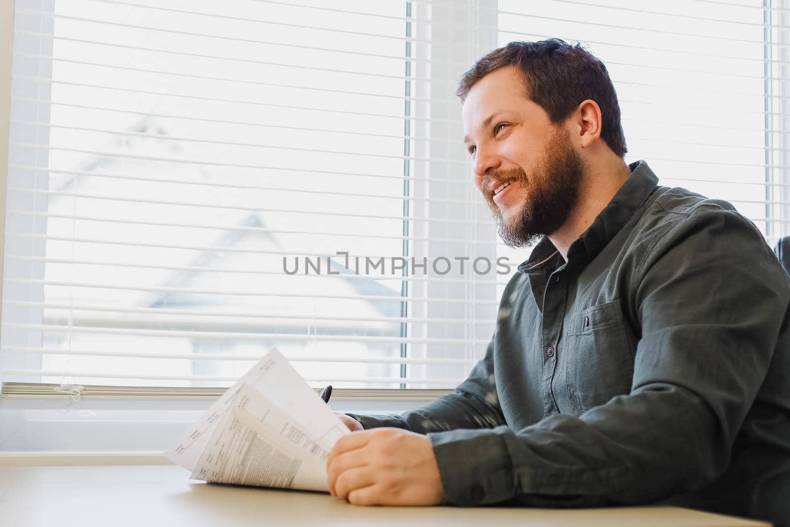 Smiling business director writing report at cabinet near window. Concept of paper work, filling documents and professional occupation. Adult man wearing shirt working with papers and sitting at desk.