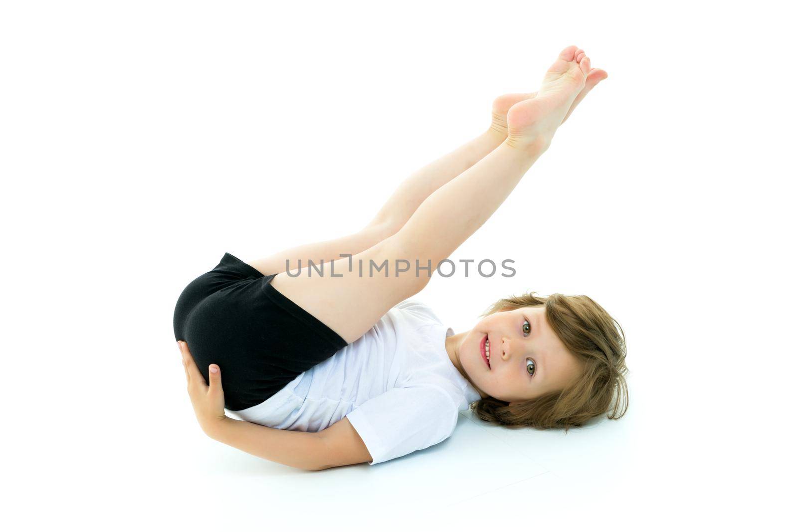 Cute little girl performs gymnastic exercises on the floor in the studio on a white background. The concept of sports, the physical development of children