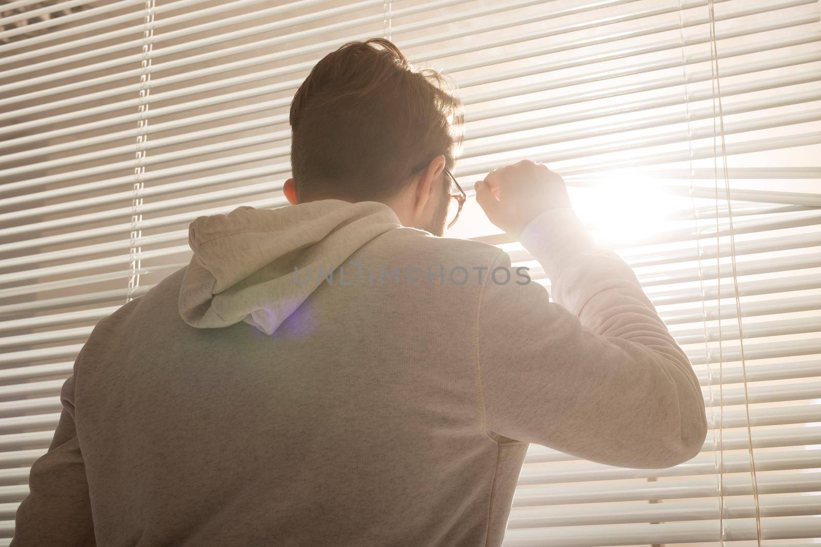 Rear view of young stylish man with beard and coffee peeking through hole in window blinds and looking out into street. Enjoying the morning sun and positivity