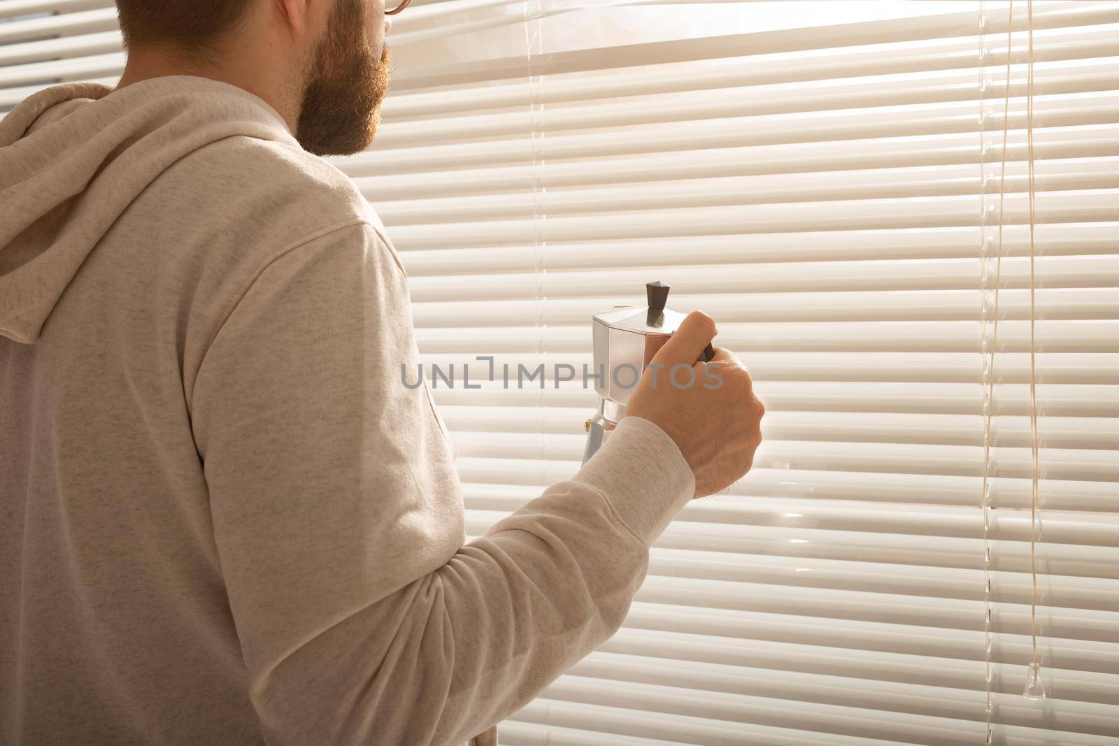 Rear view of young stylish man with beard and coffee peeking through hole in window blinds and looking out into street. Enjoying the morning sun and positivity