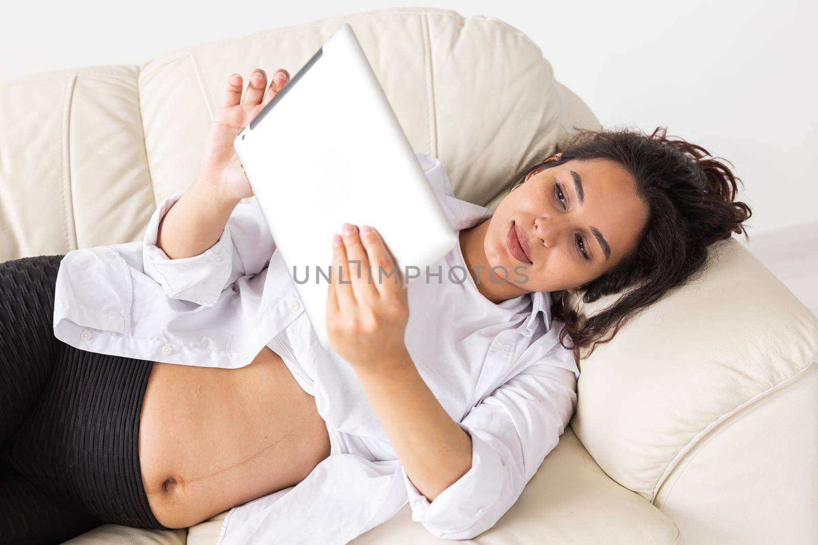 Latin pregnant woman using tablet computer sitting on sofa at home. Pregnancy and information for parenthood