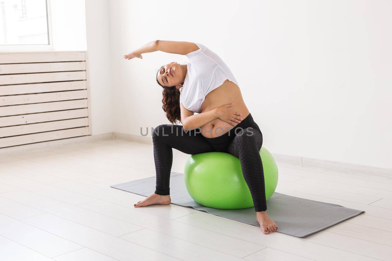 A young pregnant woman doing relaxation exercise using a fitness ball while sitting on a mat and holding her tummy