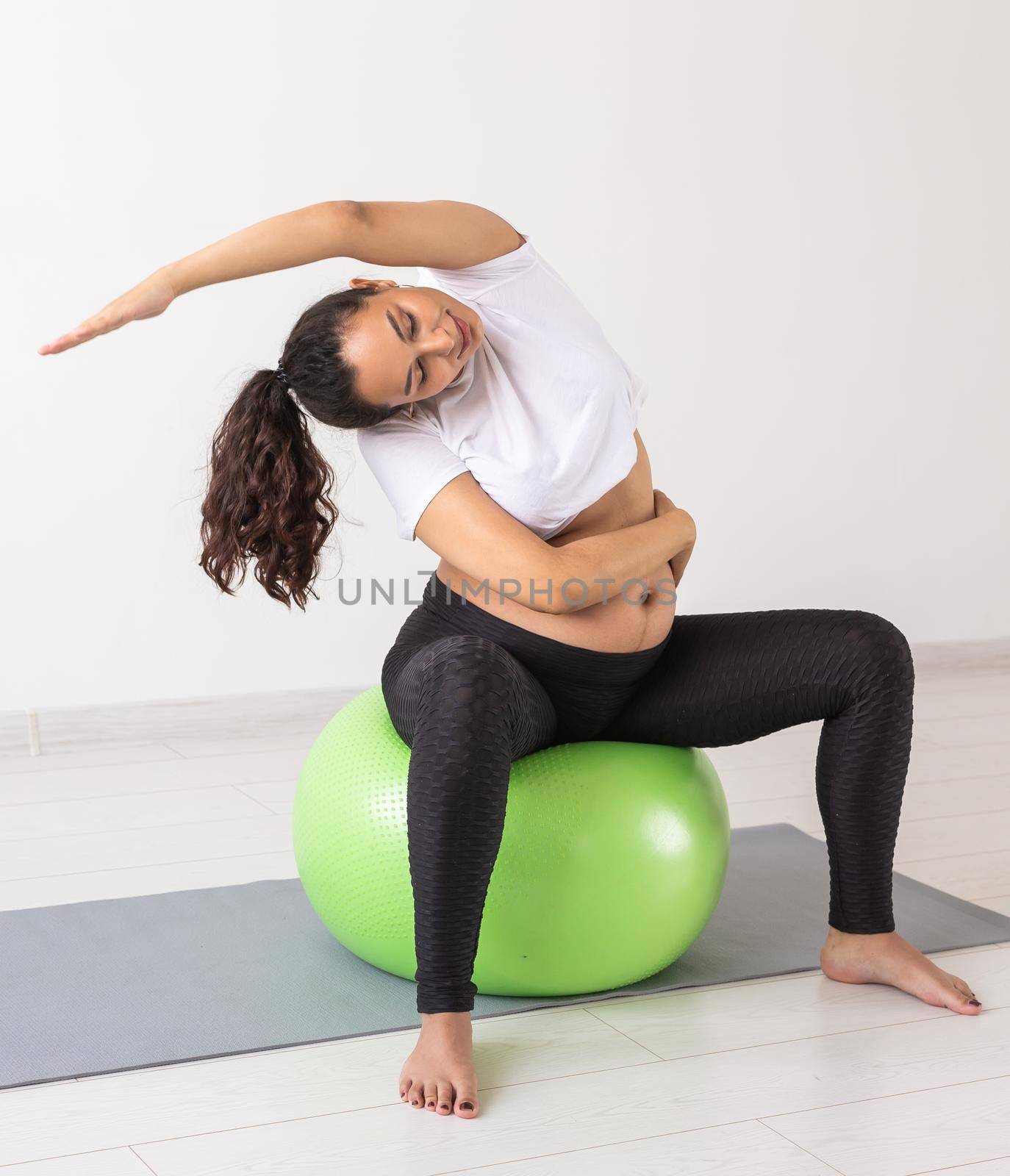 Young flexible pregnant woman doing gymnastics on rug on the floor on white background. Preparing the body for easy childbirth