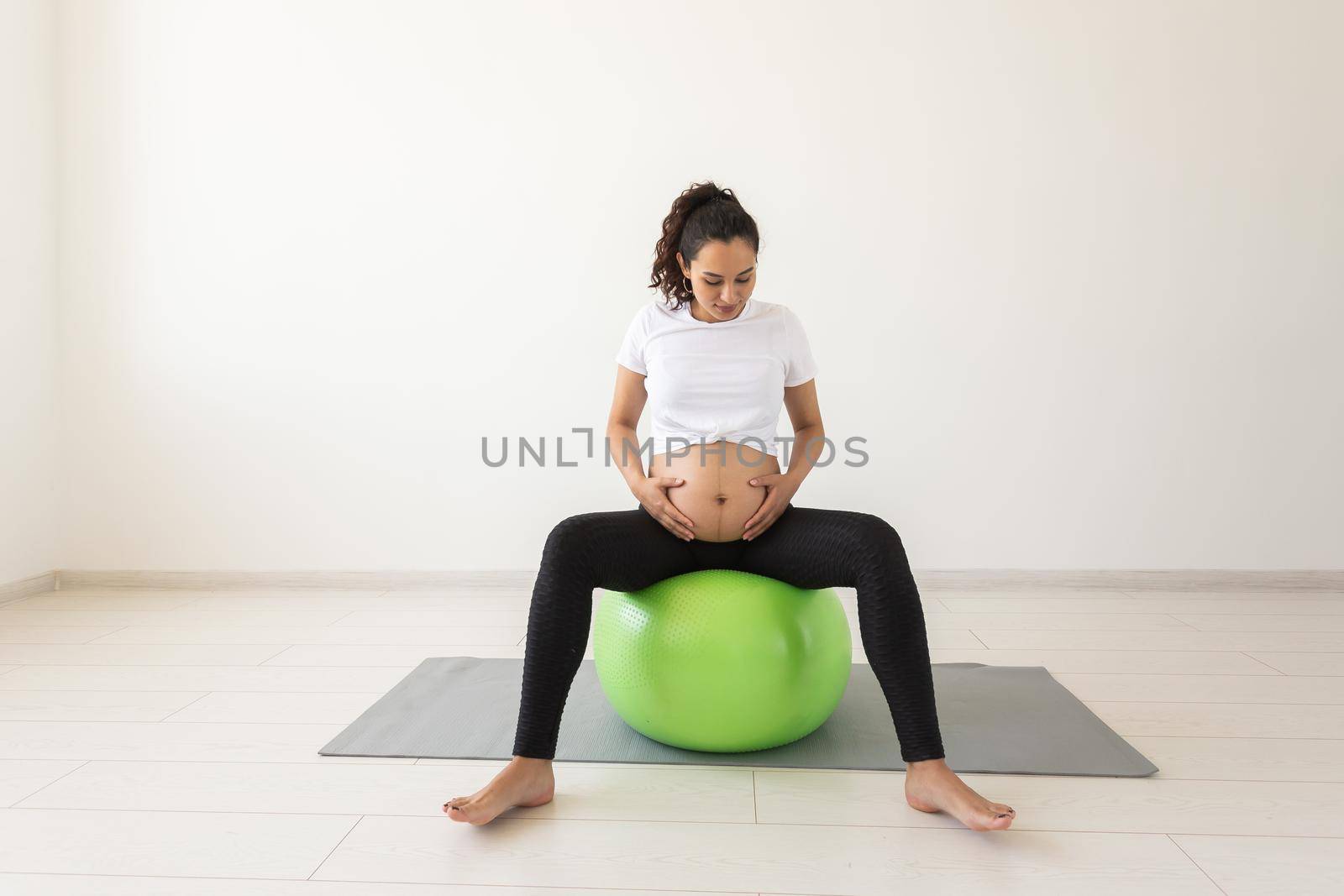 A young pregnant woman doing relaxation exercise using a fitness ball while sitting on a mat and holding her tummy