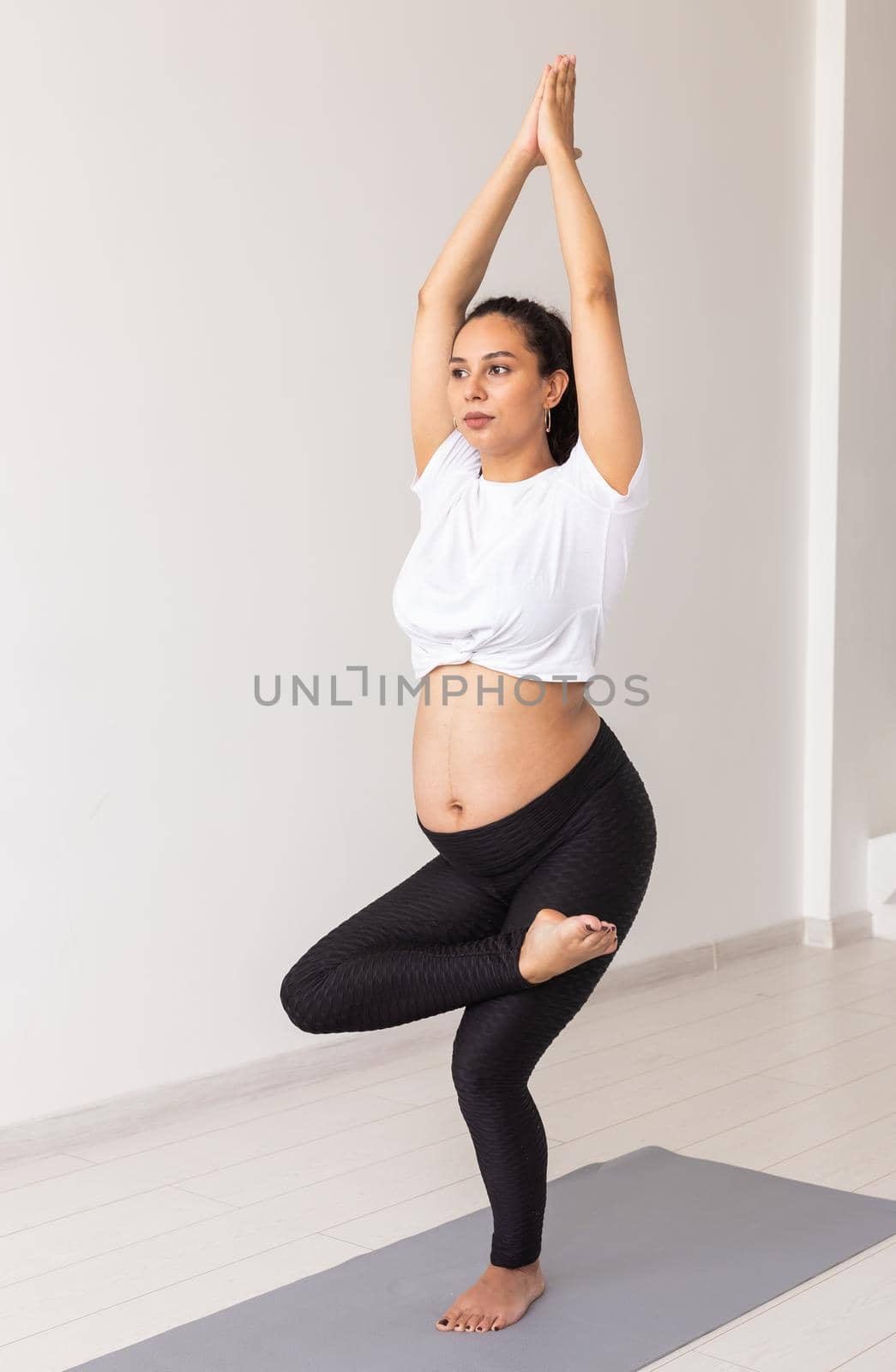 Cute flexible pregnant woman is doing gymnastics on rug on the floor on white background. Concept of preparing the body for a healthy childbirth.