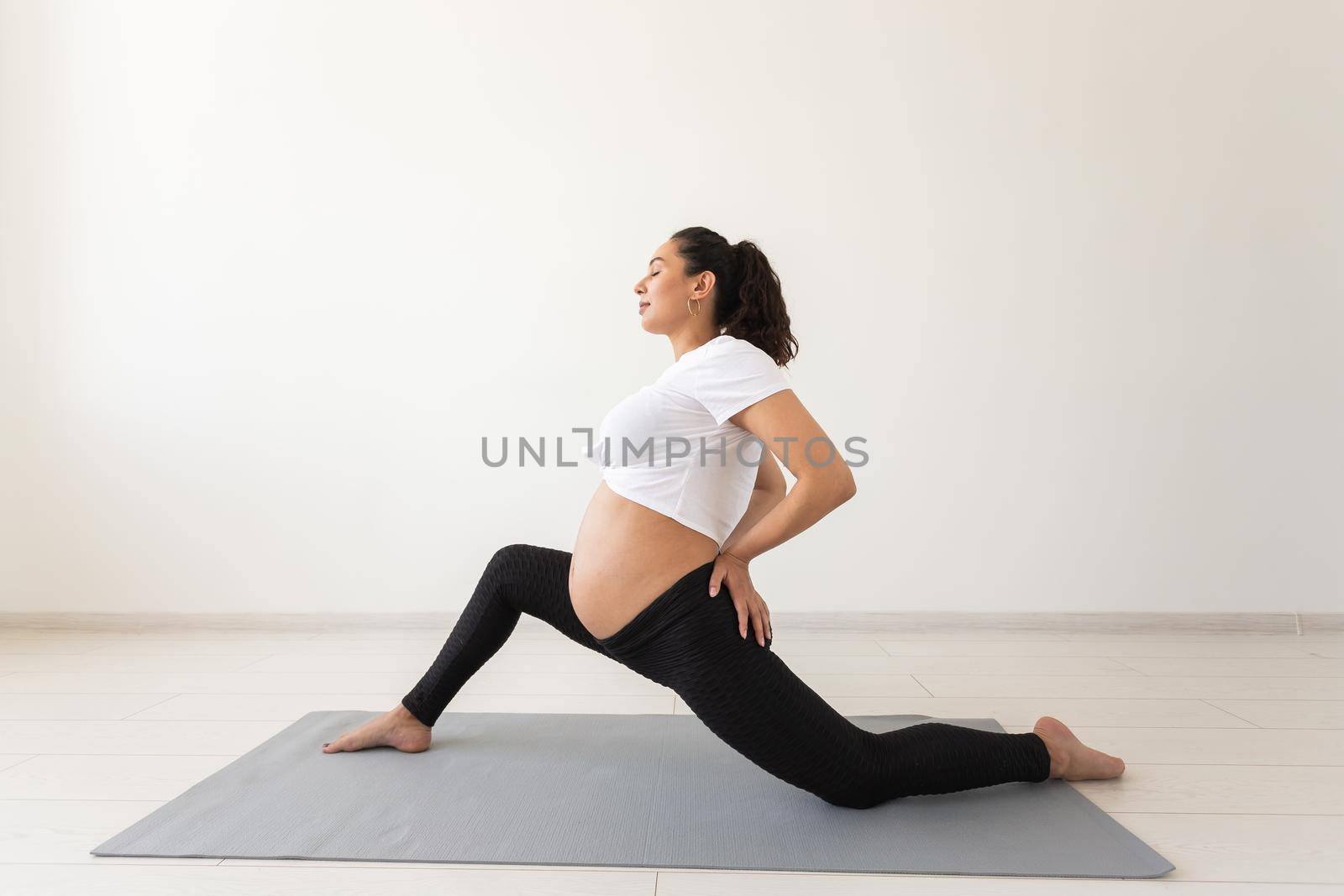 Young flexible pregnant woman doing gymnastics on rug on the floor on white background. Preparing the body for easy childbirth