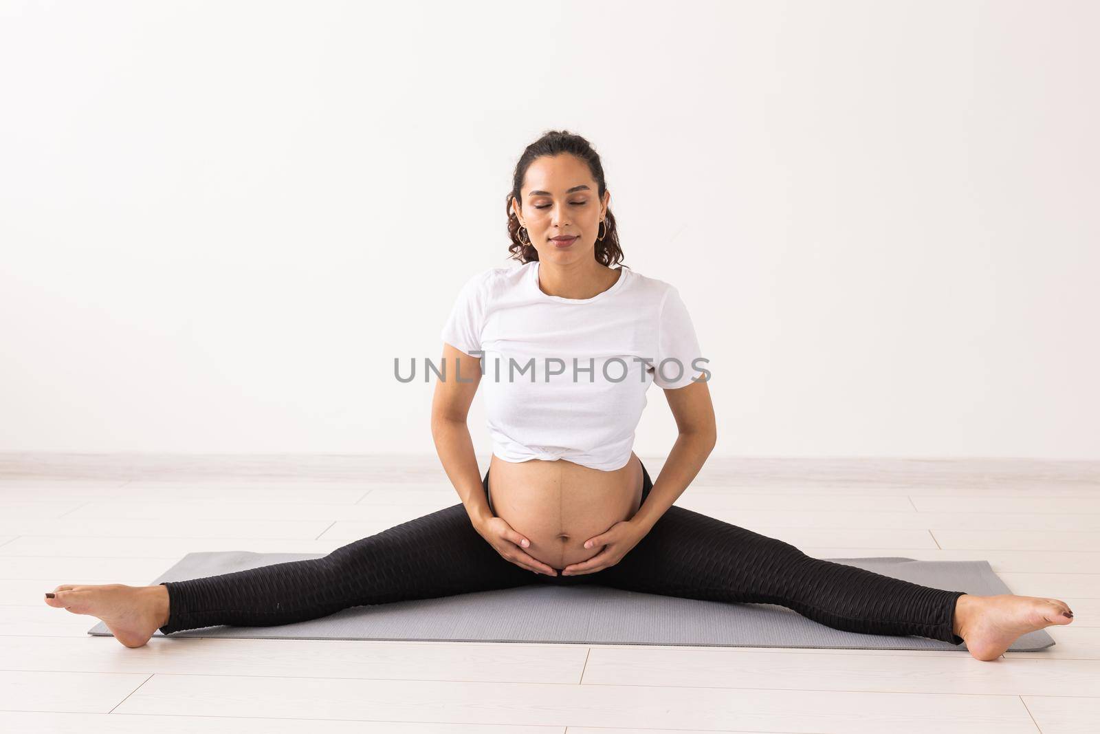 Young flexible pregnant woman doing gymnastics on rug on the floor on white background. Preparing the body for easy childbirth