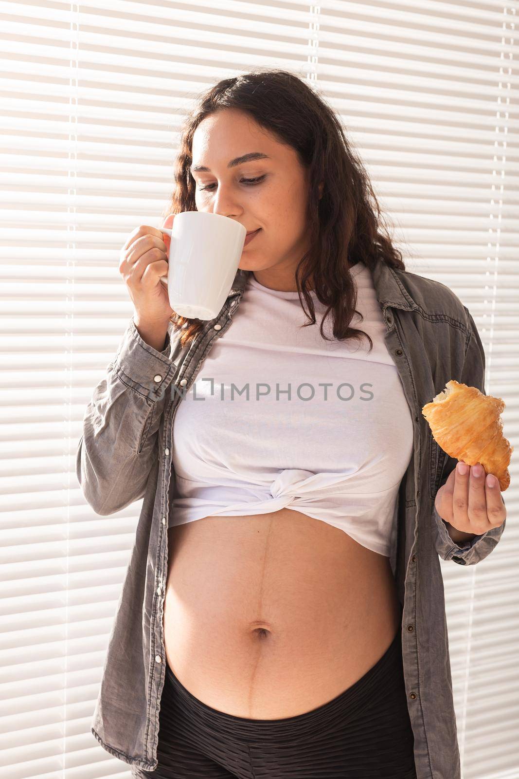Healthy beautiful pregnant woman drinking tea and eating croissant during lunch. High-calorie nutrition while waiting for birth of baby