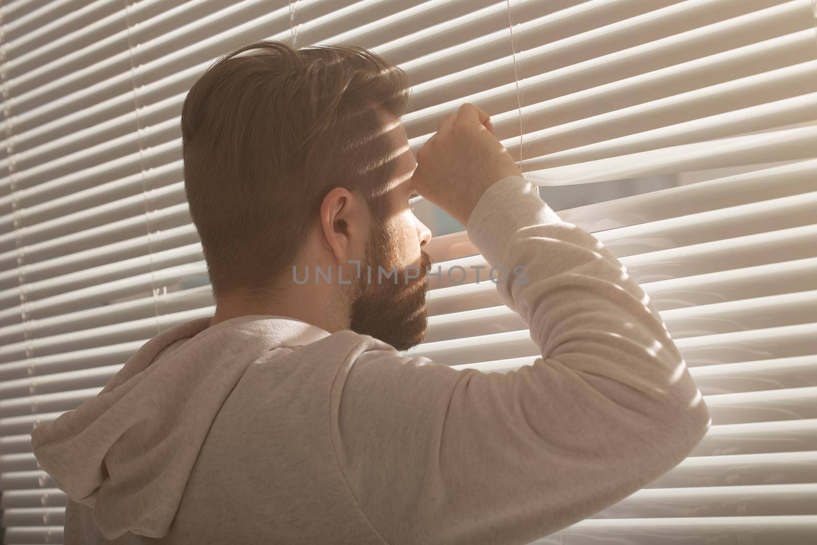 Rear view of young man with beard peeks through hole in the window blinds and looks out into the street. Surveillance and curiosity