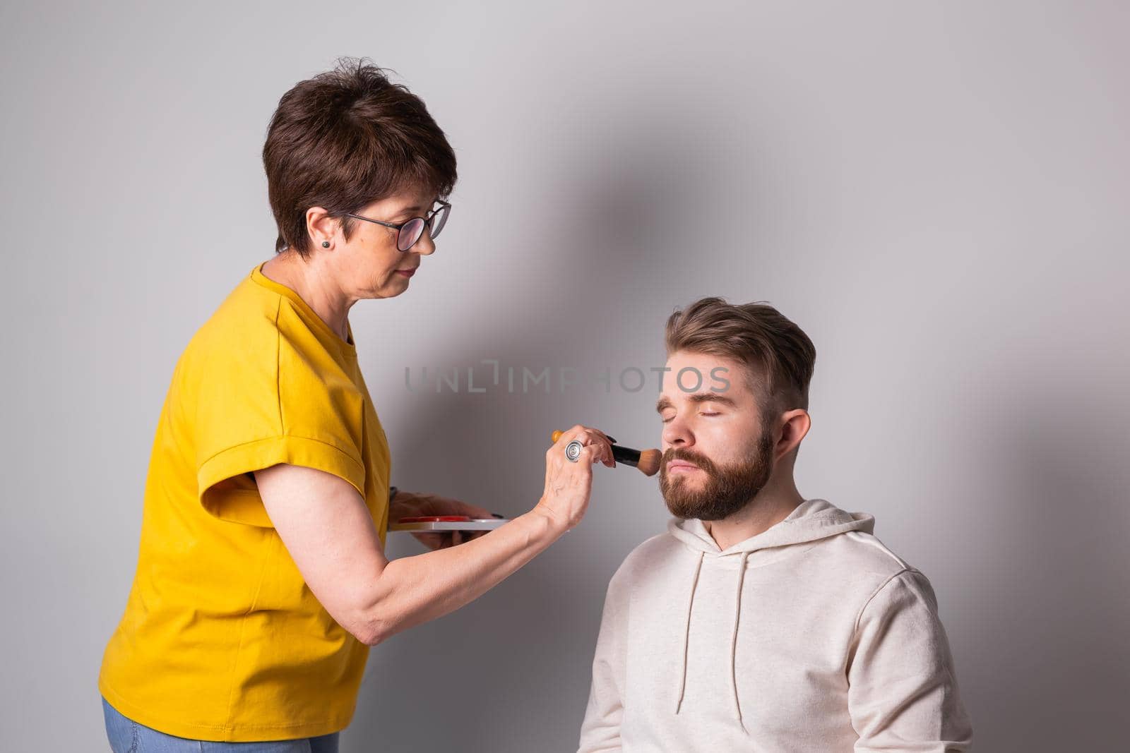 Bearded man getting makeup. Woman visagist works with brush