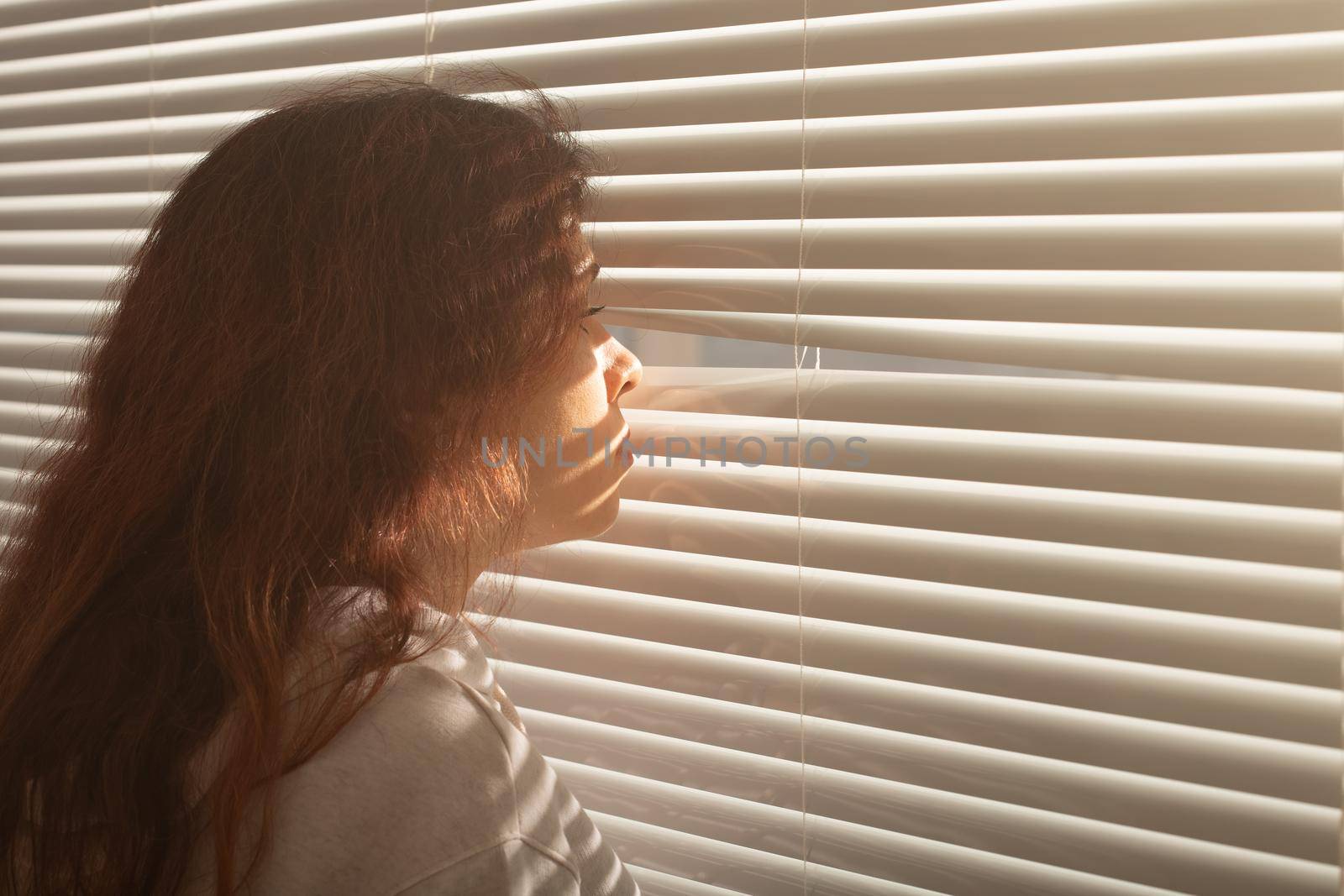 Rear view of beautiful young woman with long hair peeks through hole in the window blinds and looks out the window. Surveillance and curiosity