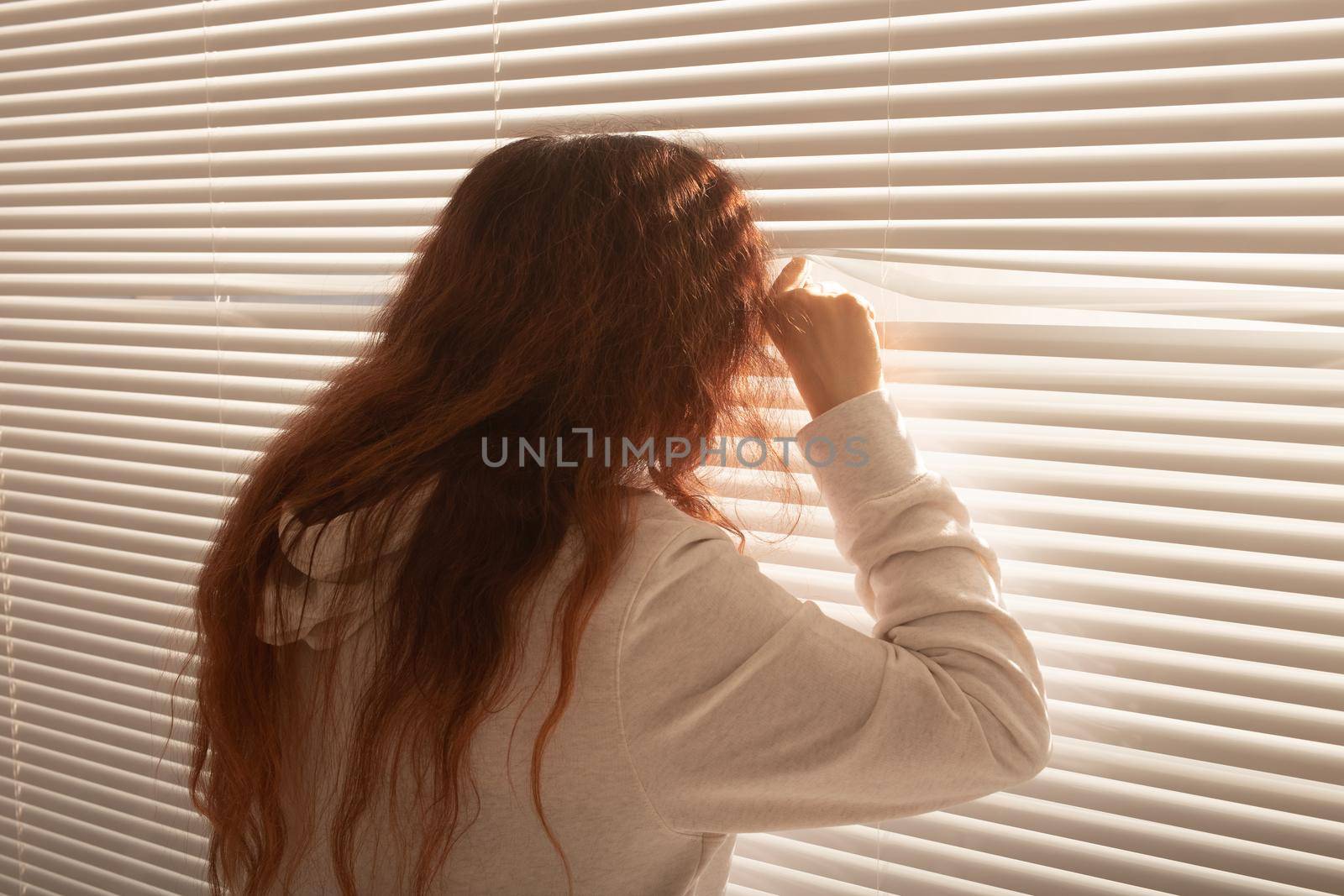 Rear view of beautiful young woman with long hair peeks through hole in the window blinds and looks out the window. Surveillance and curiosity