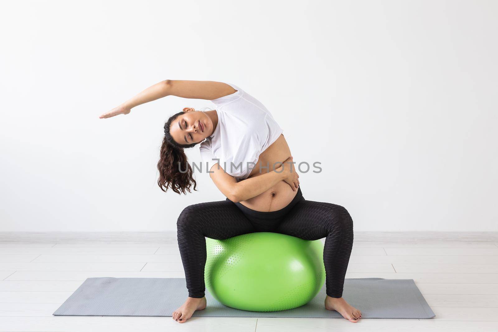 Young flexible pregnant woman doing gymnastics on rug on the floor on white background. Preparing the body for easy childbirth