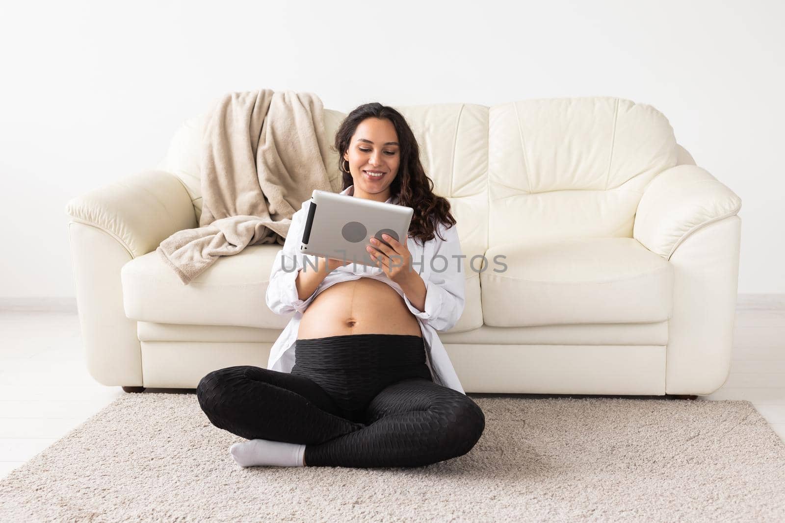 Pregnant woman holding tablet sitting on a carpet near a couch in the living room at home.