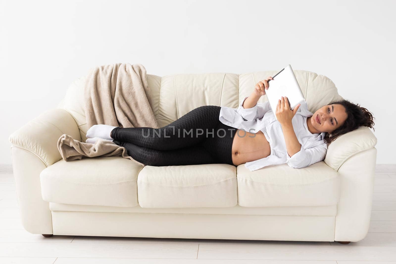 Pregnant woman holding tablet sitting on a carpet near a couch in the living room at home.