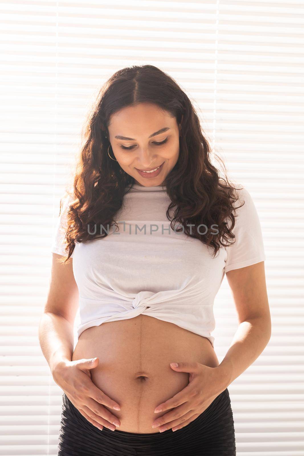 Smiling young beautiful pregnant woman touching her belly and rejoicing. Concept of positive and pleasant feelings while waiting for the baby by Satura86