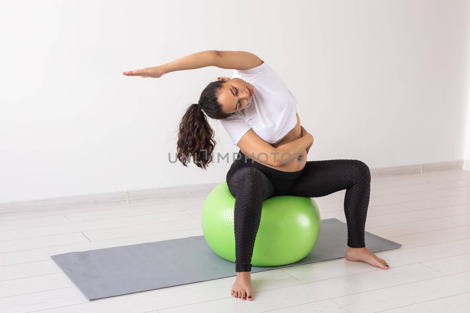 Young flexible pregnant woman doing gymnastics on rug on the floor on white background. Preparing the body for easy childbirth