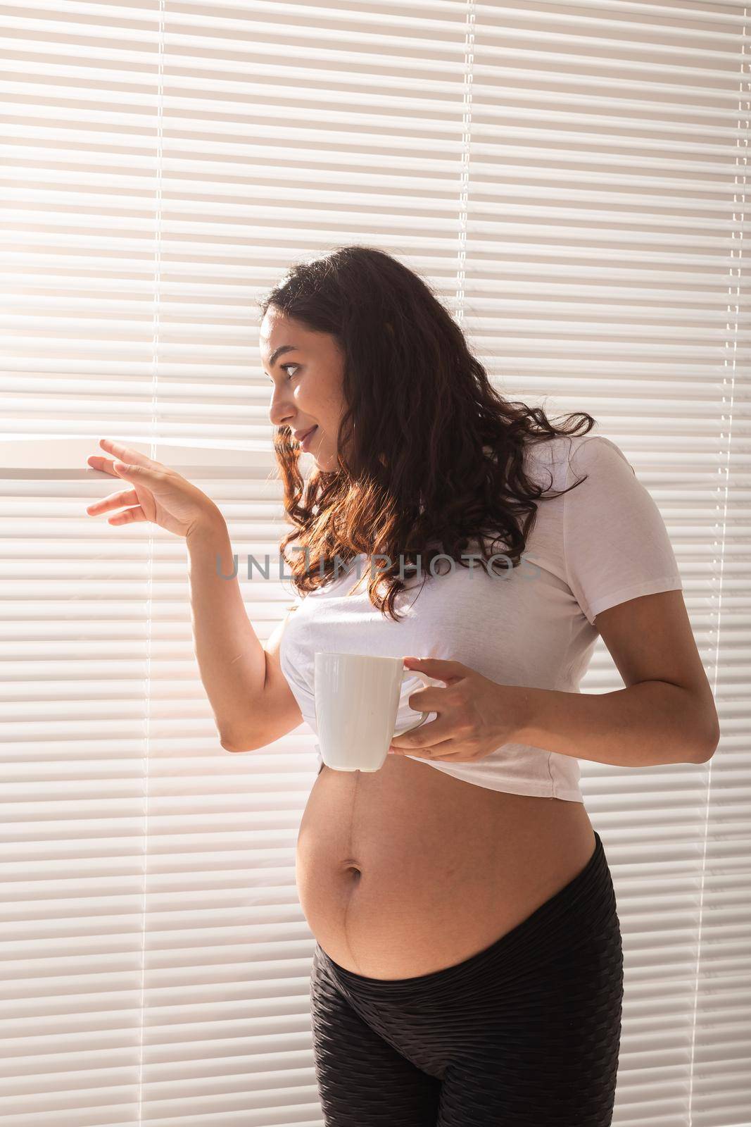 Curious young beautiful pregnant woman drinking tea and looking through the blinds at the window. Concept of joy and good news while waiting for baby by Satura86