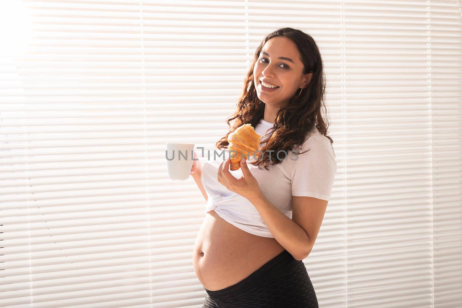 Beautiful pregnant woman holding croissant and cup of coffee in her hands during morning breakfast. Good health and positive attitude while expecting baby