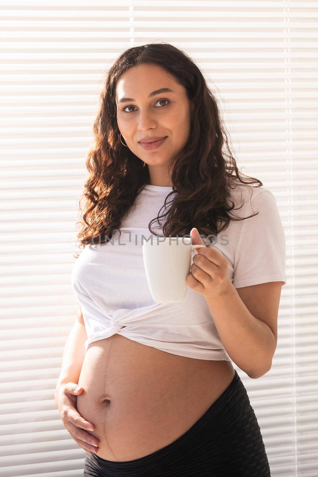 Smiling young beautiful pregnant woman touching her belly and rejoicing. Concept of positive and pleasant feelings while waiting for the baby.