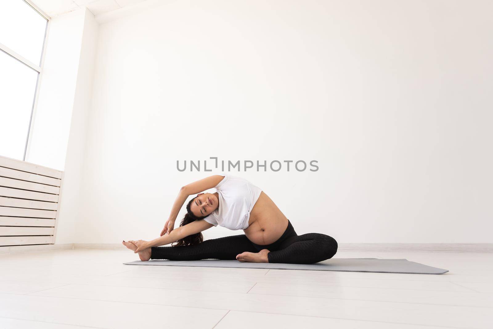 Young flexible pregnant woman doing gymnastics on rug on the floor on white background. Preparing the body for easy childbirth