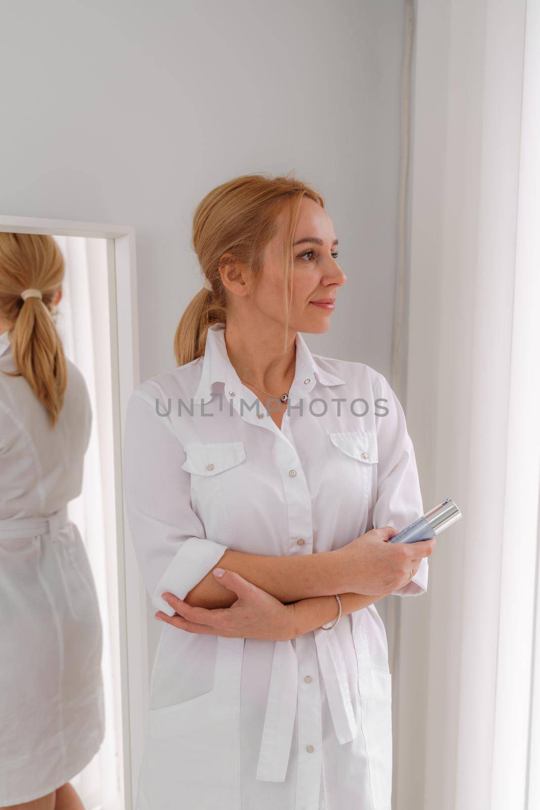 A blonde woman in white formal clothes looks out the window and is reflected in the mirror. On a white background