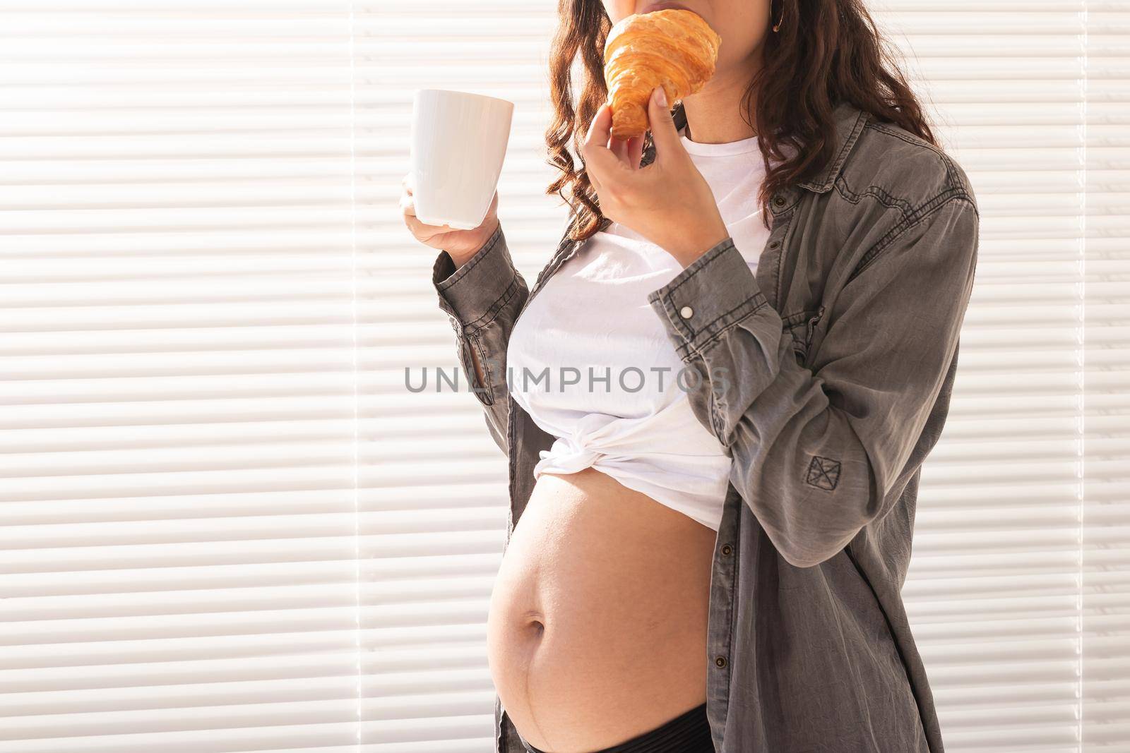 Healthy beautiful pregnant woman drinking tea and eating croissant during lunch. High-calorie nutrition while waiting for birth of baby