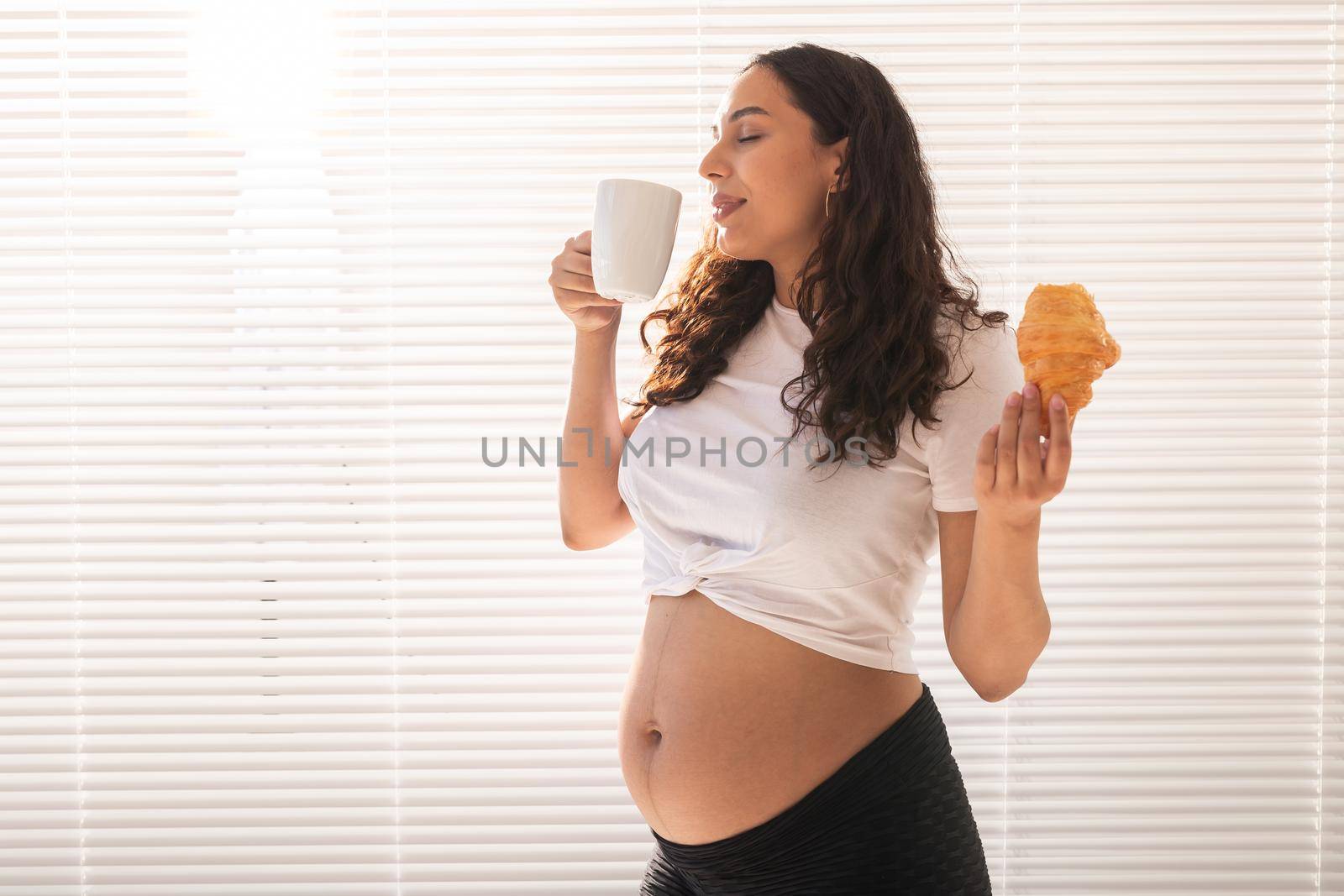 Pregnant woman eating croissant and drinks coffee. Pregnancy and maternity leave.