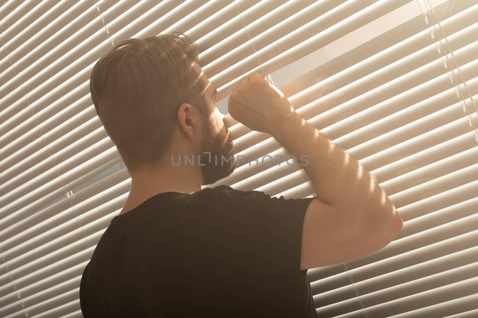 Rear view of young man with beard peeks through hole in the window blinds and looks out into the street. Surveillance and curiosity