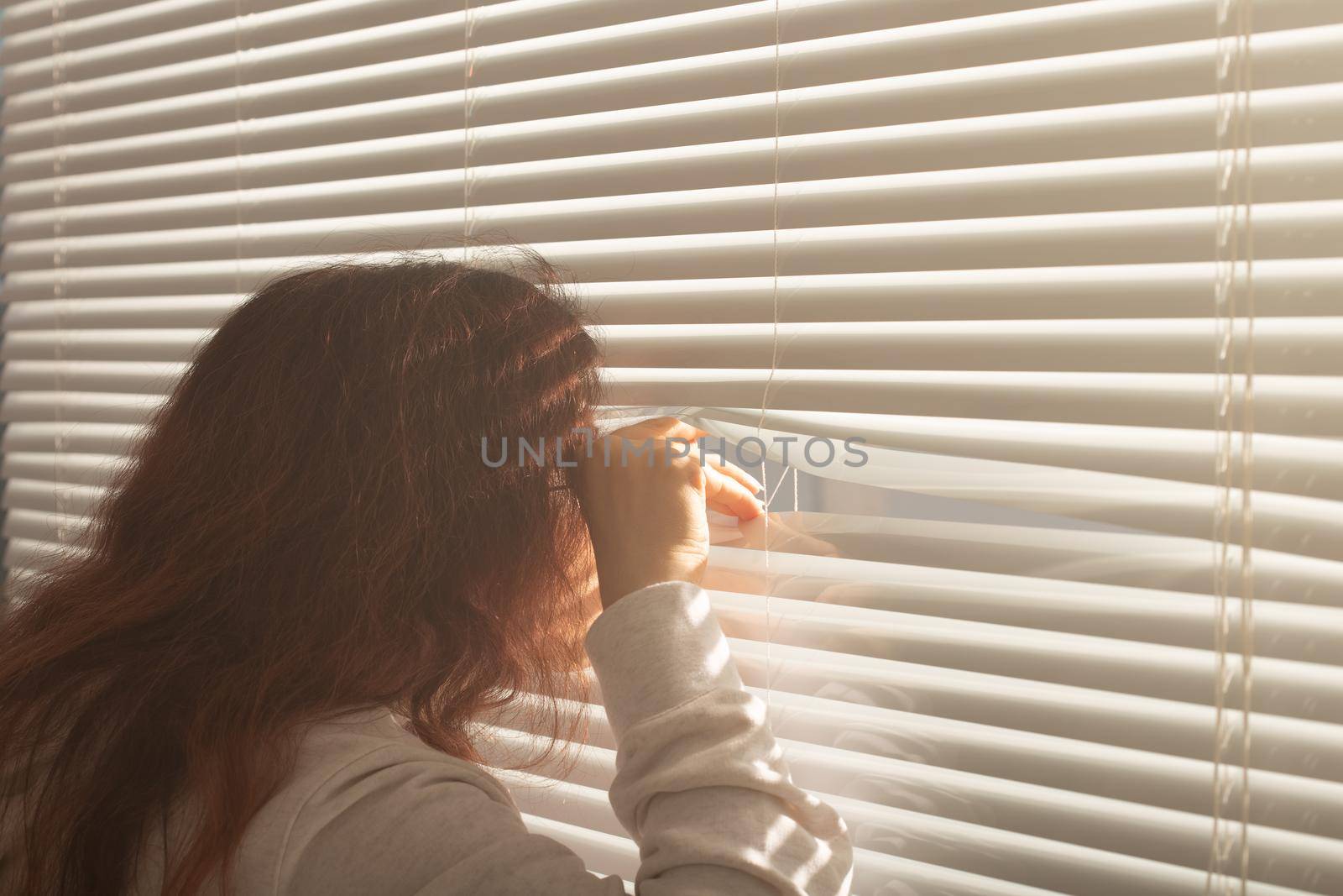 Rear view of beautiful young woman with long hair peeks through hole in the window blinds and looks out the window. Surveillance and curiosity
