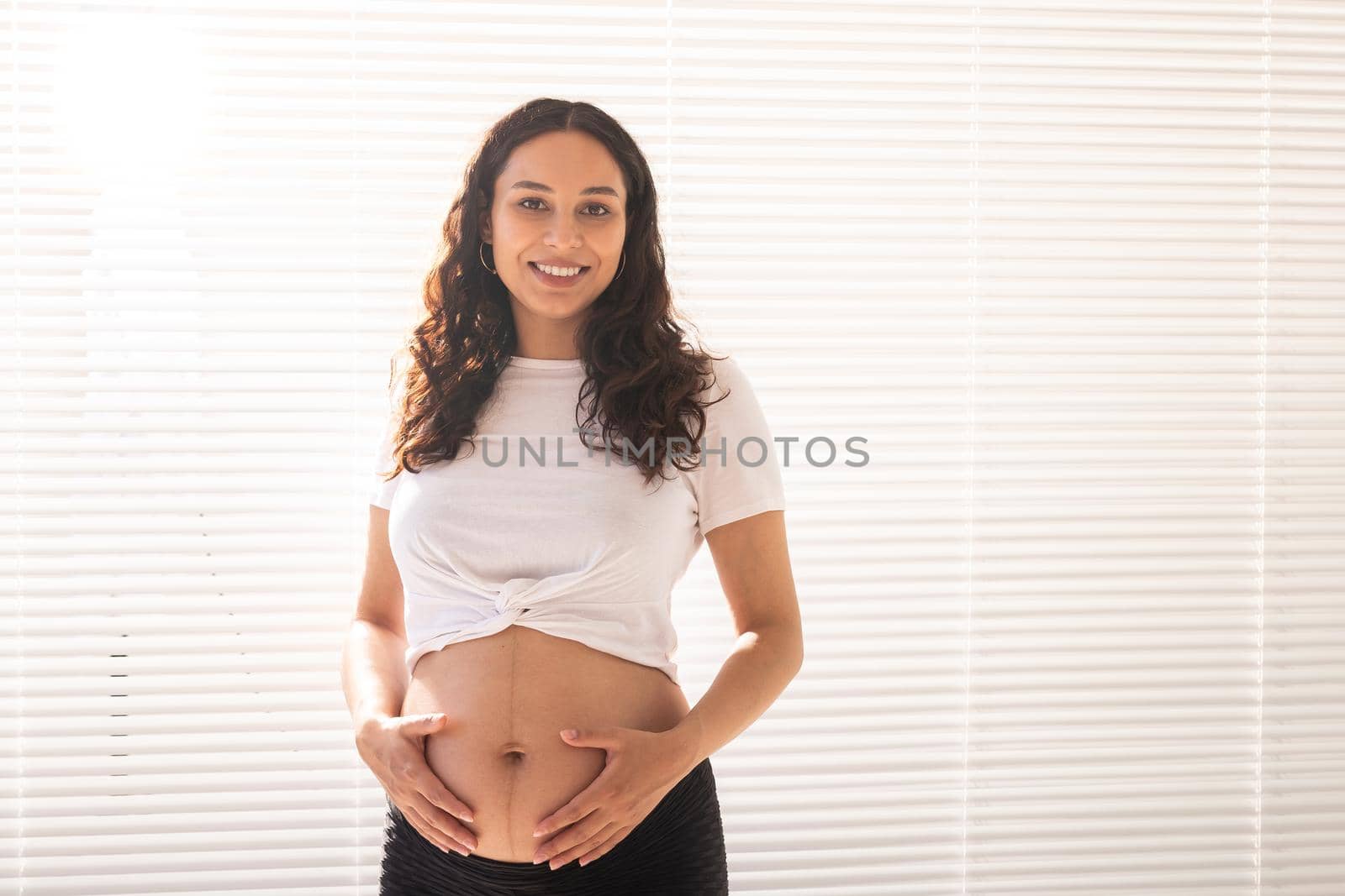 Smiling young beautiful pregnant woman touching her belly and rejoicing. Concept of positive and pleasant feelings while waiting for the baby.