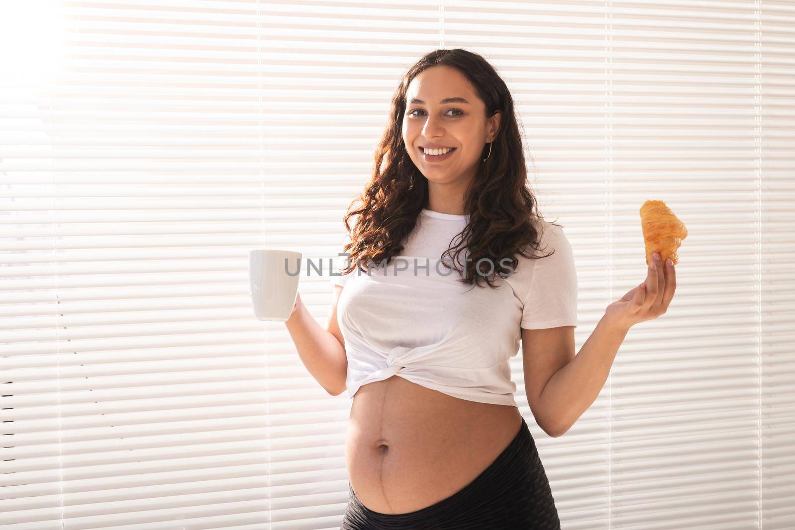 Beautiful pregnant woman holding croissant and cup of coffee in her hands during morning breakfast. Good health and positive attitude while expecting baby
