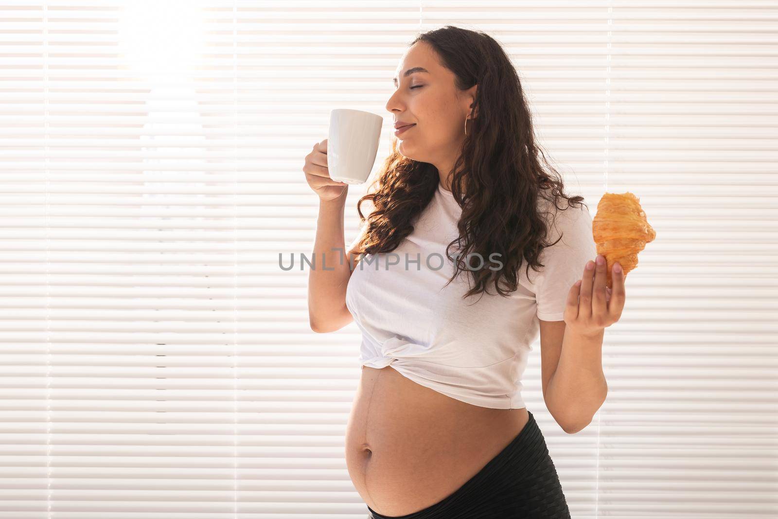 Beautiful pregnant woman holding croissant and cup of coffee in her hands during morning breakfast. Good health and positive attitude while expecting baby