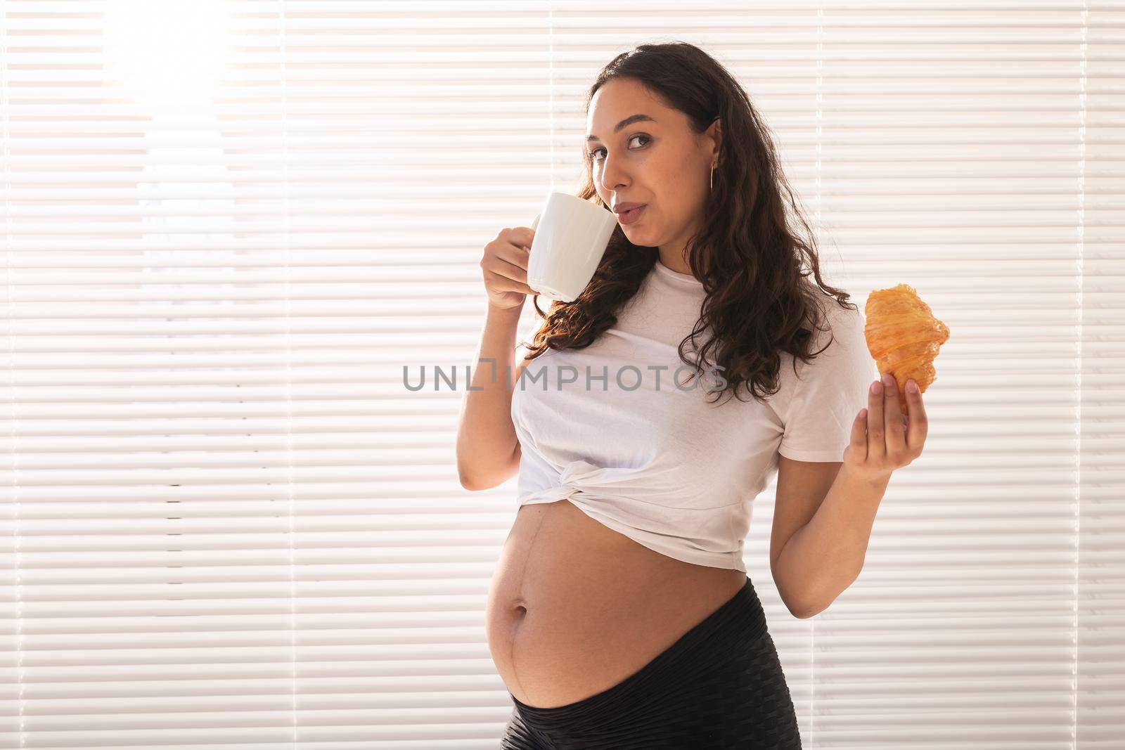 Pregnant woman eating croissant and drinks coffee. Pregnancy and maternity leave.