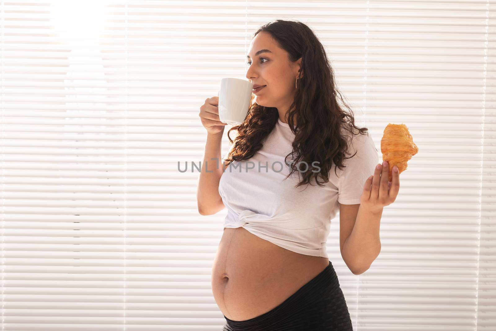 Beautiful pregnant woman holding croissant and cup of coffee in her hands during morning breakfast. Good health and positive attitude while expecting baby