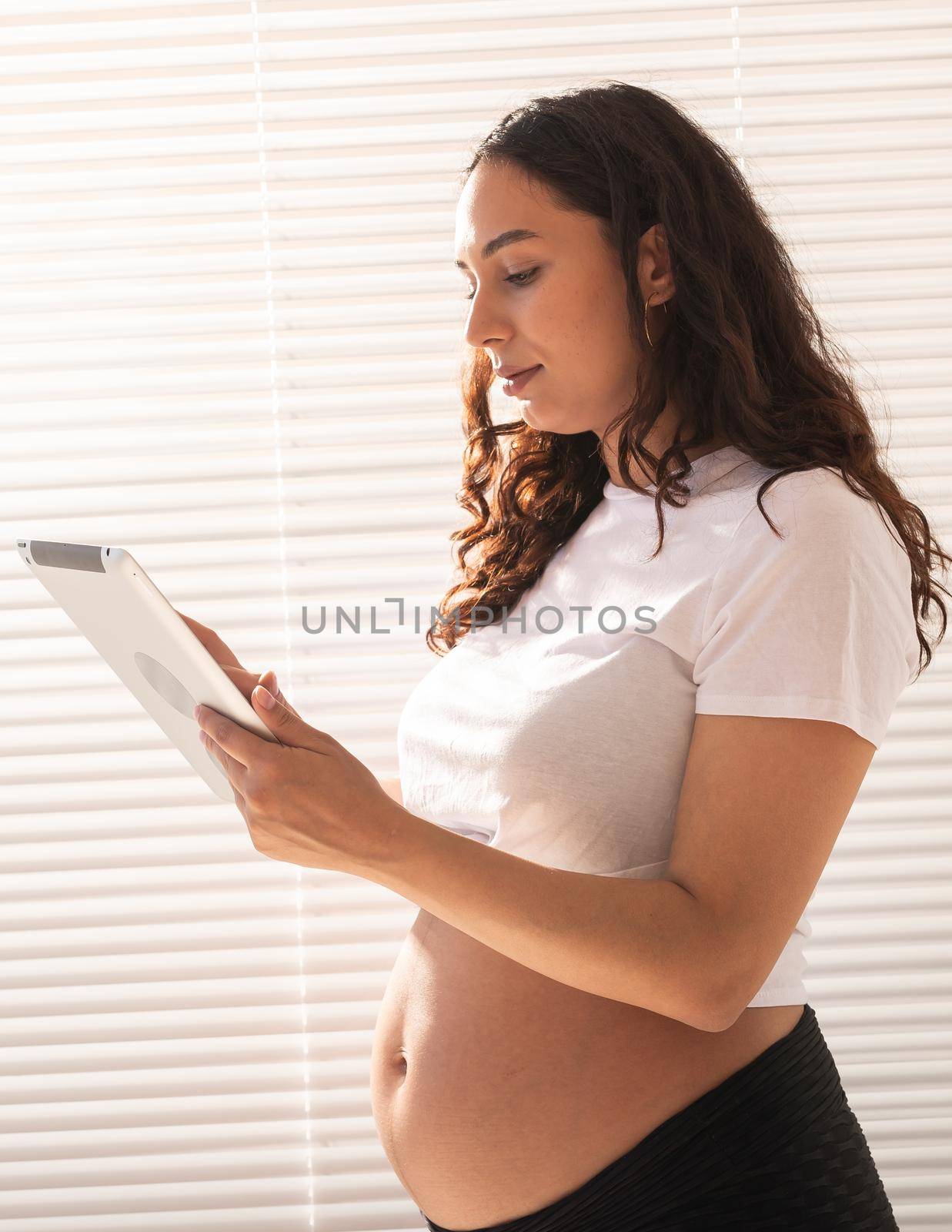 Happy pregnant young beautiful woman talking to her husband using video connection and tablet. Concept of communication and positive attitude during pregnancy by Satura86