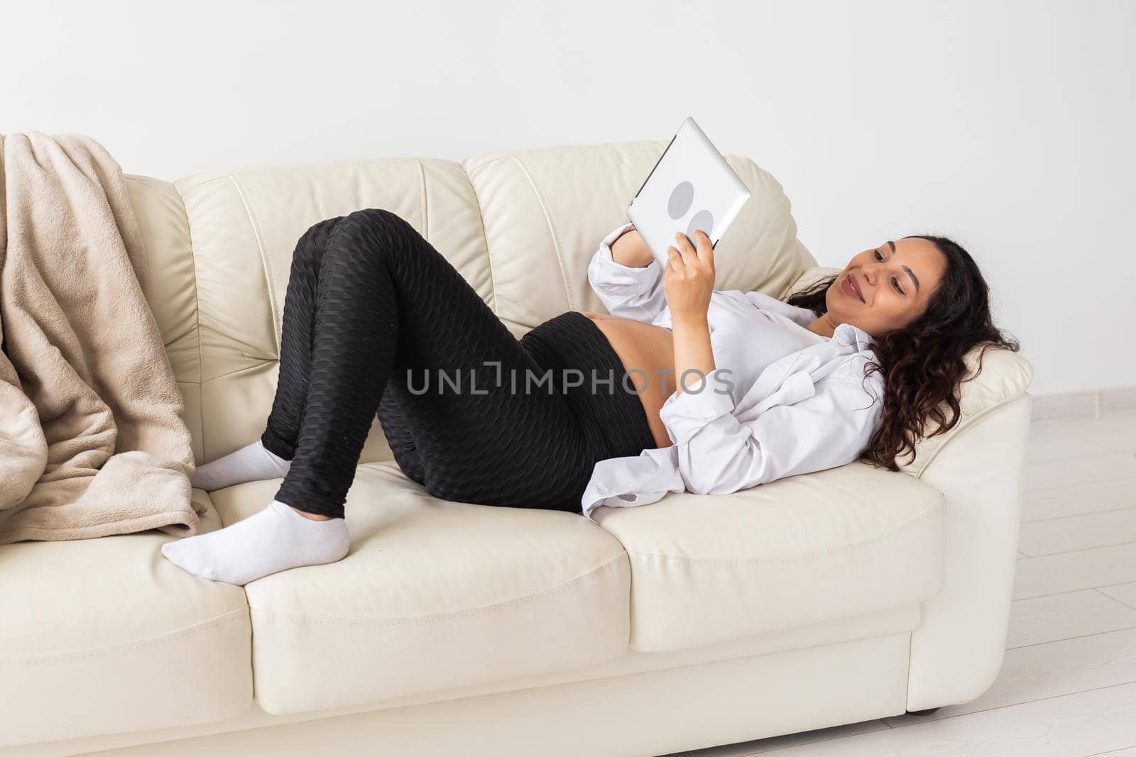Pregnant woman holding tablet sitting on a carpet near a couch in the living room at home.