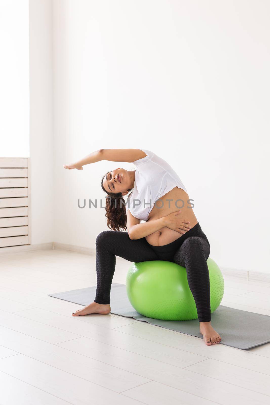 A young pregnant woman doing exercise using a fitness ball while sitting on a mat. by Satura86